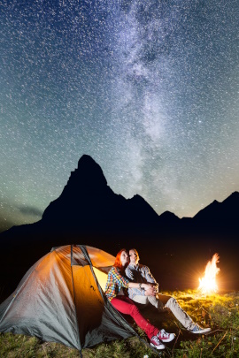 Paar sitzt nachts vorm Zelt am Lagerfeuer und betrachtet den Sternenhimmel