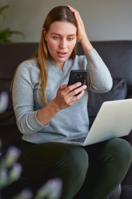 Frau mit Laptop schaut zuhause schockiert aufs Handy