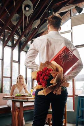 Mann überrascht Frau im Restaurant mit Geschenken hinterm Rücken