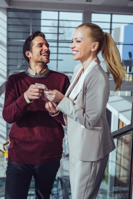 Zwei Kollegen stehen lachend im Bürogebäude und flirten