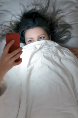 Frau liegt zugedeckt im Bett und schaut aufs Handy
