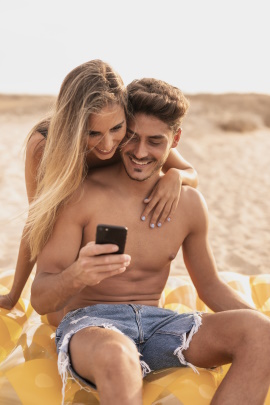 Frau umarmt Mann am Strand und schaut aufs Handy in seiner Hand