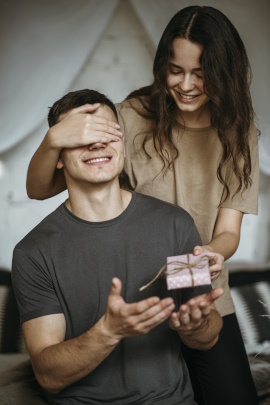 Frau hält Partner die Augen zu und überreicht ihm ein Geschenk