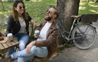 Paar sitzt lachend im Park am Tisch und trinkt Kaffee aus Bechern