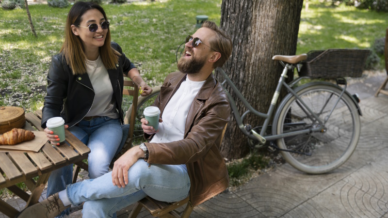 Paar sitzt lachend im Park am Tisch und trinkt Kaffee aus Bechern