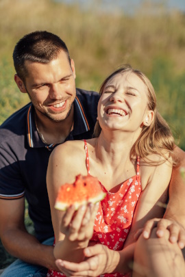 Frau sitzt mit Partner lachend im Gras und isst Wassermelone