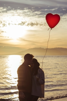 Mann und Frau schauen sich verliebt den Sonnenuntergang an und halten einen Herzballon