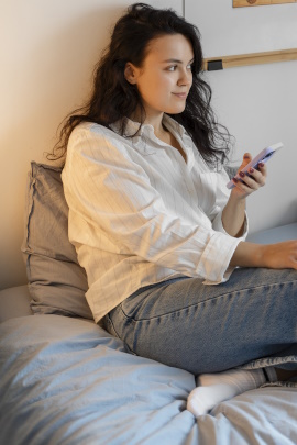 Frau mit Handy sitzt nachdenklich auf dem Bett