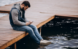 Mann sitzt einsam auf dem Steg am Wasser und schaut aufs Handy