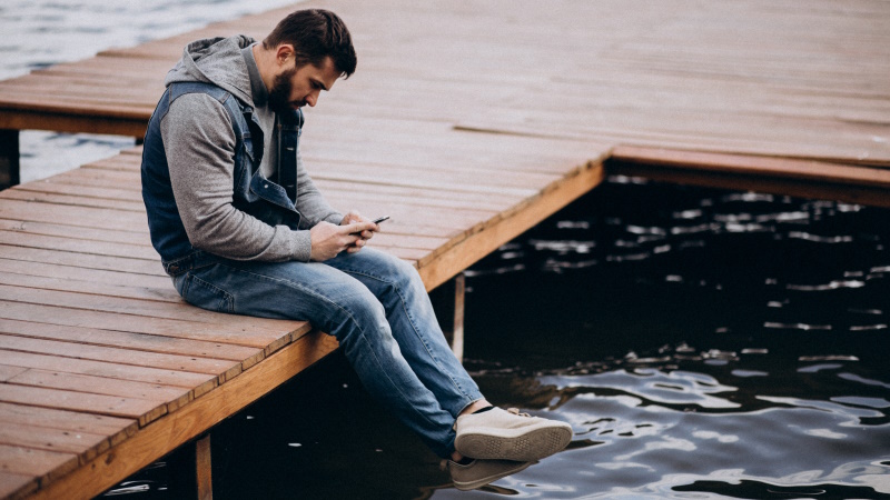 Mann sitzt einsam auf dem Steg am Wasser und schaut aufs Handy