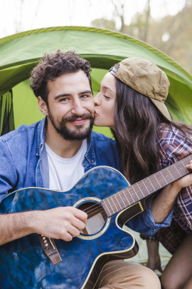 Frau küsst Gitarre spielenden Mann vor Camping-Zelt auf die Wange