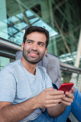 Mann sitzt lächelnd am Glasgebäude und schreibt Nachricht am Handy