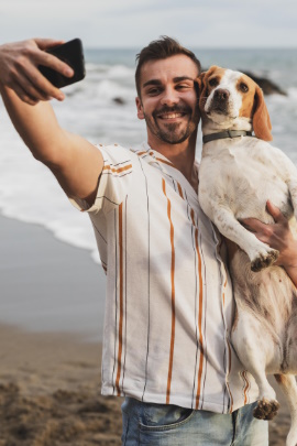 Mann steht am Strand und macht ein Selfie mit Hund im Arm