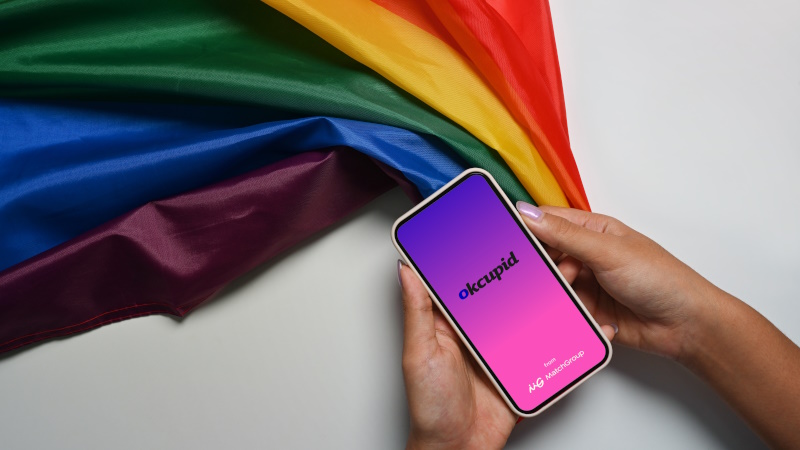 Frau mit Regenbogenfahne hält Handy mit OkCupid-App-Startbildschirm