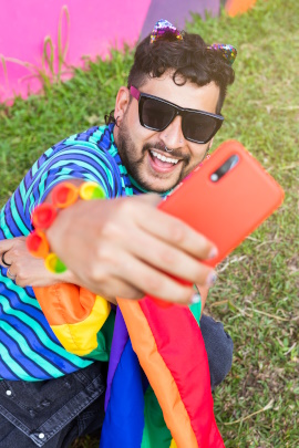 Mann mit Sonnenbrille und Regenbogenfahne macht Selfie im Gras liegend
