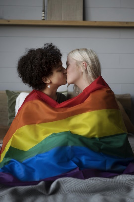Zwei Frauen mit Regenbogenfahne sitzen küssend auf dem Bett