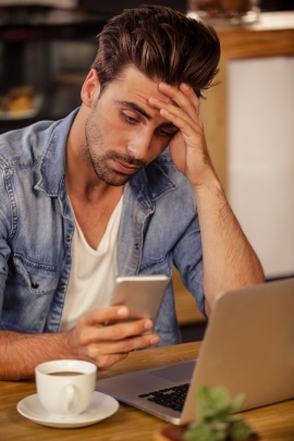 Mann mit Handy und Kaffee sitzt frustriert am Laptop