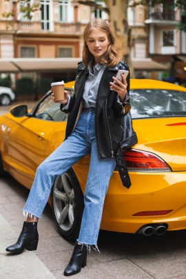 Frau mit Kaffeebecher lehnt am gelben Sportwagen und schaut aufs Handy