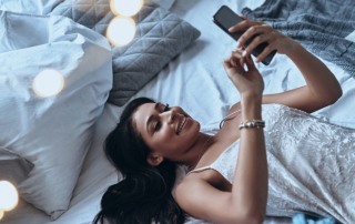 Lächelnde Frau im Nachthemd liegt auf dem Bett und liest Nachricht am Handy