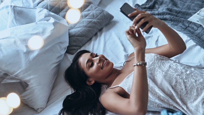 Lächelnde Frau im Nachthemd liegt auf dem Bett und liest Nachricht am Handy