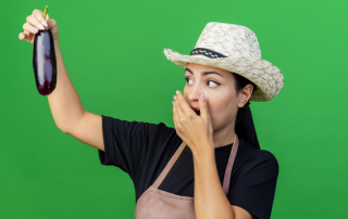 Frau mit Hut blickt schockiert auf Aubergine in ihrer Hand