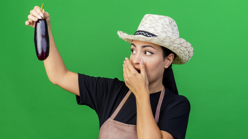 Frau mit Hut blickt schockiert auf Aubergine in ihrer Hand