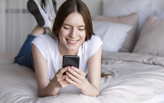 Frau liegt auf dem Bett und schaut lächelnd aufs Smartphone