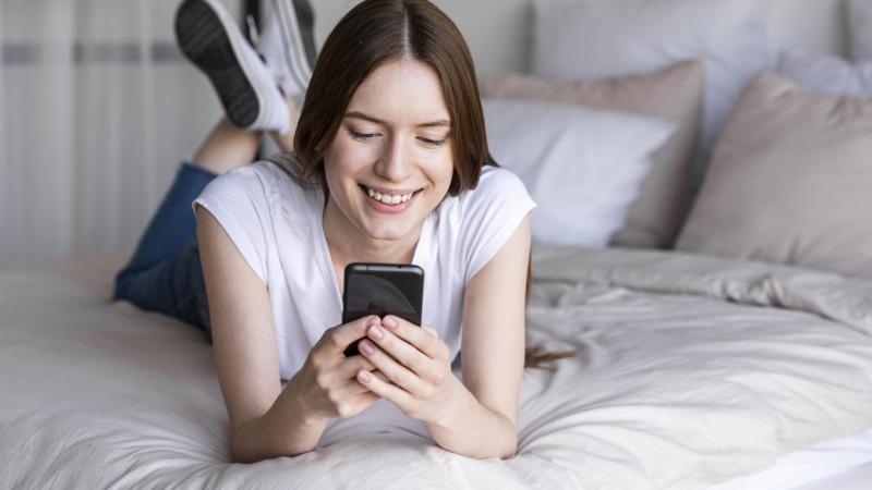 Frau liegt auf dem Bett und schaut lächelnd aufs Smartphone