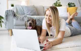 Lächelnde Frau sitzt mit Hund vorm Laptop im Wohnzimmer