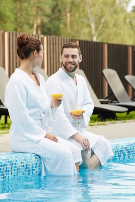 Paar im Bademantel sitzt draußen am Schwimmbeckenrand und trinkt Saft