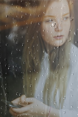 Frau mit Smartphone schaut bei Regen aus dem Fenster