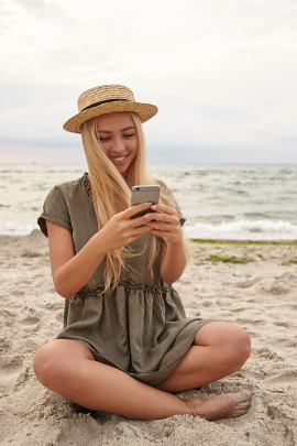 Tinder-Nutzerin sitzt am Strand und schaut lachend aufs Handy