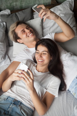 Paar mit Smartphones liegt lachend im Bett