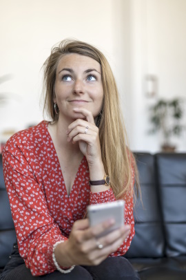 Frau mit Handy sitzt auf dem Sofa und schaut nachdenklich nach oben