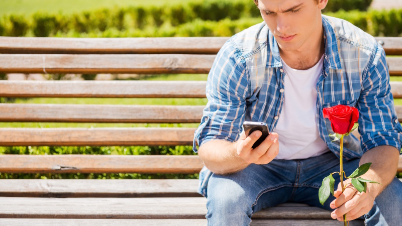 Mann sitzt auf Parkbank mit Smartphone und Rose in den Händen
