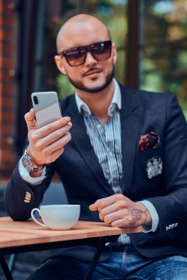 Eleganter Mann mit Glatze und Sonnenbrille sitzt mit Handy im Café