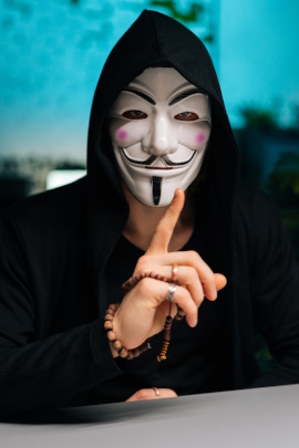 Hacker mit Maske sitzt am Tisch mit erhobenem Zeigefinger