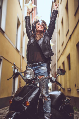 Frau steht lachend auf dem Motorroller und streckt die Arme in die Luft